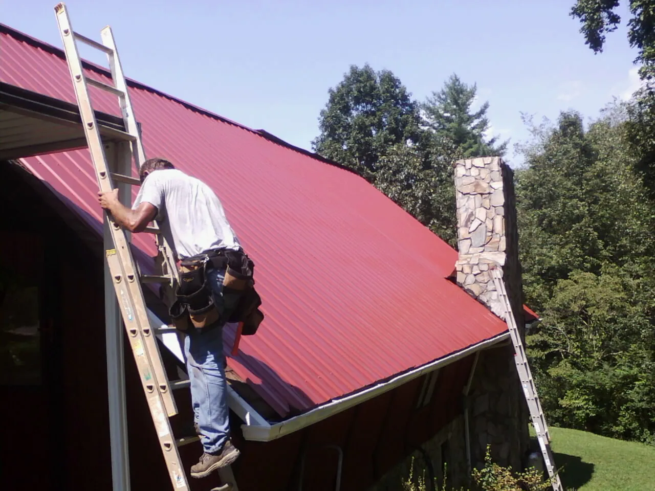 Roofer on a ladder beside a red metal roof and stone chimney — Soffit & Fascia Repair by Roofing Brothers in Ripon